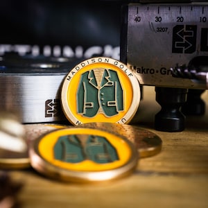 May include: Close-up of a gold-coloured golf ball marker with a green blazer design. The text "MADDISON GOLF" is printed around the edge of the marker.