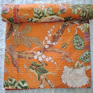 Könnte beinhalten: Orangefarbene Kantha-Steppdecke mit Blumenmuster. Die Decke ist aus Baumwolle und hat ein handgesticktes Design. Die Decke ist aufgerollt und hat einen braunen und orangen Rand.