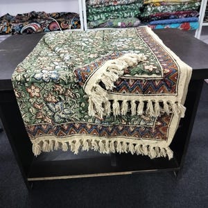 Peut inclure: Un chemin de table à motifs verts et bruns avec une frange blanche. Le chemin de table présente un motif floral et une bordure géométrique.