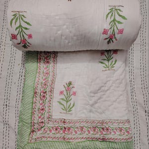 May include: White quilted blanket with a floral design in pink and green. The blanket has a rolled-up section at the top, displaying the floral pattern. The border features a green and white striped pattern, adding a decorative touch to the bedding.