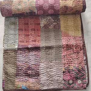 May include: A patchwork quilt featuring a variety of rectangular fabric panels in shades of brown, pink, tan, and olive green. The quilt is partially rolled at the top and displays intricate white running stitch patterns across the entire surface.