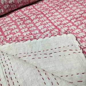 Puede incluir: Una colcha de algodón rosa con estampado de elefantes blancos. La colcha está doblada por la mitad, mostrando las costuras en la parte posterior.