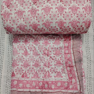 Puede incluir: Una colcha de algodón rosa y blanco con un patrón floral. La colcha está doblada por la mitad y tiene un borde marrón.