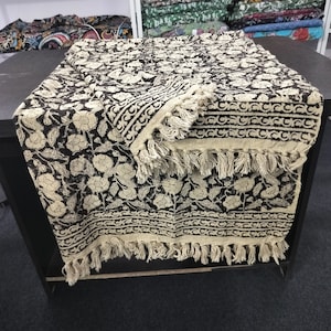 May include: A black and white cotton throw blanket with a floral pattern and fringe trim. The blanket is folded in half and has a textured surface.
