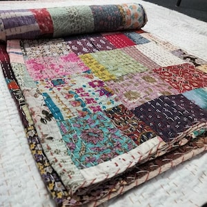 Op de afbeelding: Een patchwork quilt met een opgerold gedeelte. De quilt heeft een verscheidenheid aan bloemen- en patroonstoffen in roze, blauw, groen en rood. De quilt is met de hand gestikt met witte draad, waardoor een textuur ontstaat.