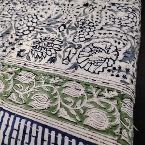 Op de afbeelding: Witte stof met een blauwe en groene bloemenprint. De stof heeft een herhalend patroon van bloemen en bladeren. De stof is zacht en comfortabel.