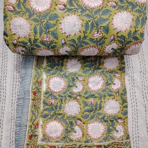 Op de afbeelding: Een groene en witte quilt met een bloemenpatroon en een rand van rode en witte bloemen. De quilt is opgerold en de stof is zichtbaar.