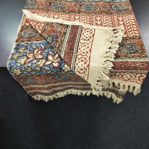 Könnte beinhalten: Eine cremefarbene Wolldecke mit einem Paisley-Muster in Blautönen, Rot und Braun. Die Decke hat einen Fransenrand und ist in der Hälfte gefaltet.