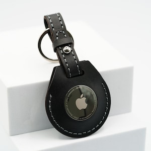 May include: A black leather keychain with a silver Apple logo and the text "Designed by Apple in California. Assembled in China. Ultra Wideband. AirTag. Bluetooth.  "