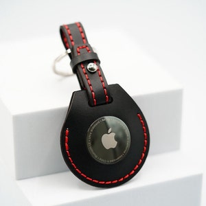 May include: Black leather keychain with a red stitched border. The keychain has a round silver tag with the Apple logo and the text "AirTag - Ultra Wideband - Designed by Apple in California - Assembled in China - Bluetooth LE".