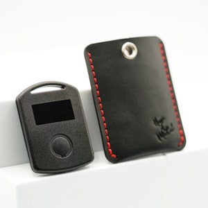 May include: A black key fob with a small screen and a button. The key fob is next to a black leather key holder with red stitching.