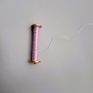 May include: A spool of pink thread with a gold metal top and bottom. The thread is unwinding from the spool.