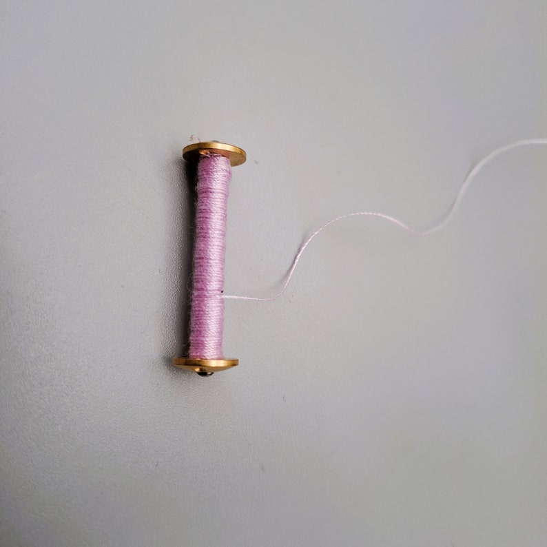 May include: A spool of pink thread with a gold metal top and bottom. The thread is unwinding from the spool.