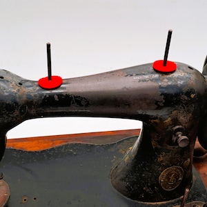 May include: An antique black sewing machine with a brown wooden base. The machine has two red spool holders and a large hand wheel. The metal has a distressed, aged appearance, suggesting it is vintage.