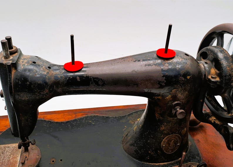 May include: An antique black sewing machine with a brown wooden base. The machine has two red spool holders and a large hand wheel. The metal has a distressed, aged appearance, suggesting it is vintage.