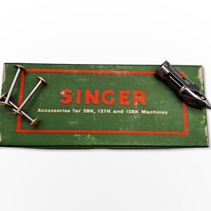 Puede incluir: Una caja verde con la palabra "SINGER" en letras rojas y el texto "Accessories for 28K, 127K and 128K Machines". La caja contiene un accesorio de m&aacute;quina de coser plateado y cuatro carretes plateados con centros dorados.