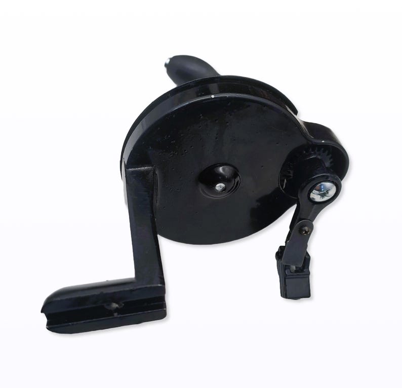 May include: A black manual pencil sharpener with a hand crank. The sharpener has a circular body with a handle and a small gear mechanism. The crank is attached to the side of the sharpener.