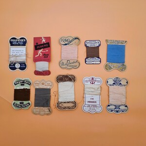 May include: A collection of vintage thread spools in various colors, including pink, blue, brown, and cream. Each spool has a unique label with text and decorative elements. The spools are arranged on an orange background.