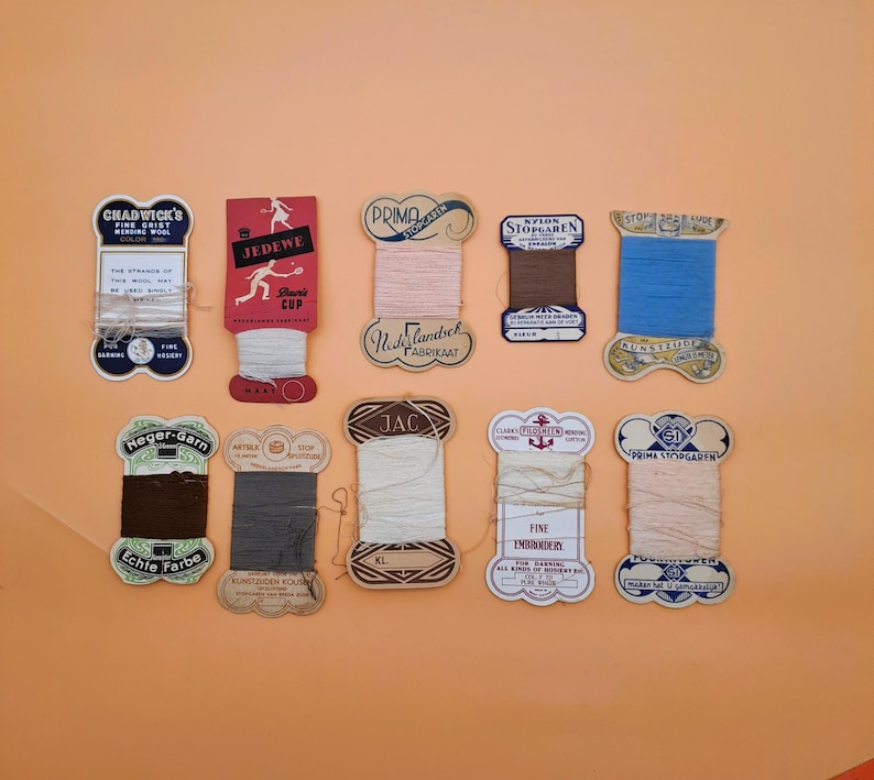 May include: A collection of vintage thread spools in various colors, including pink, blue, brown, and cream. Each spool has a unique label with text and decorative elements. The spools are arranged on an orange background.