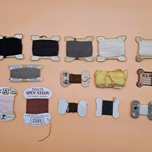May include: A collection of vintage thread spools in various colors, including black, gray, white, and brown. Some spools have labels with text, and the background is a solid peach color. The spools are arranged on a flat surface.