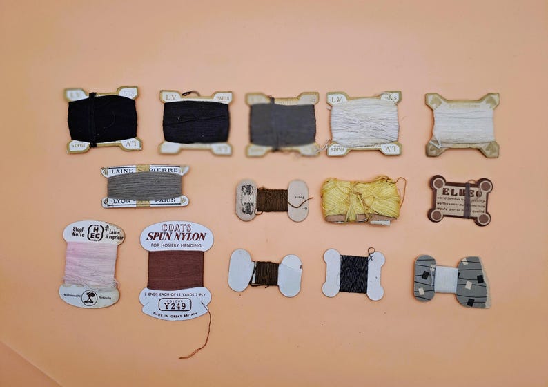 May include: A collection of vintage thread spools in various colors, including black, gray, white, and brown. Some spools have labels with text, and the background is a solid peach color. The spools are arranged on a flat surface.