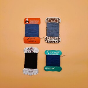 May include: Four spools of thread in various colors: dark blue, light blue, and black. Each spool has a different label with text, including "MODINETJE MAASWOL", "JAC", "NYLON", and "FLORTISS". The spools are arranged on a peach-colored background.