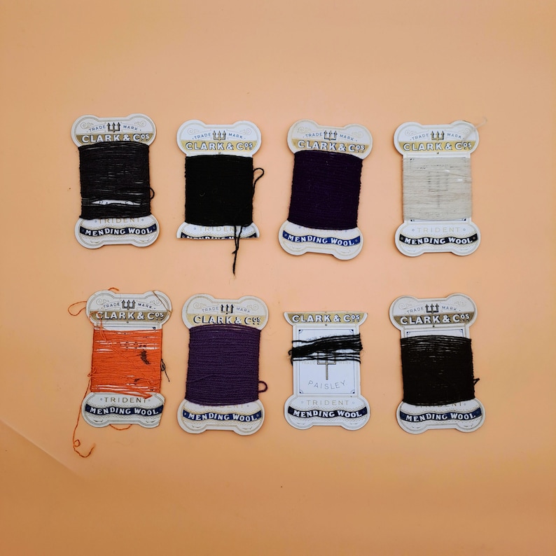 May include: A collection of eight spools of mending wool in various colors, including black, purple, orange, and beige. Each spool is labeled with "CLARK & CO" and "MENDING WOOL" on a cream-colored label.