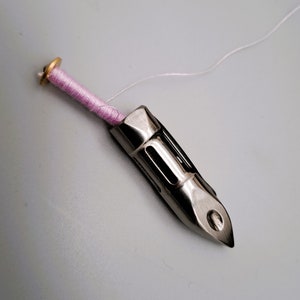 May include: A silver sewing machine bobbin winder with a pink thread wound around it. The thread is attached to a white thread.