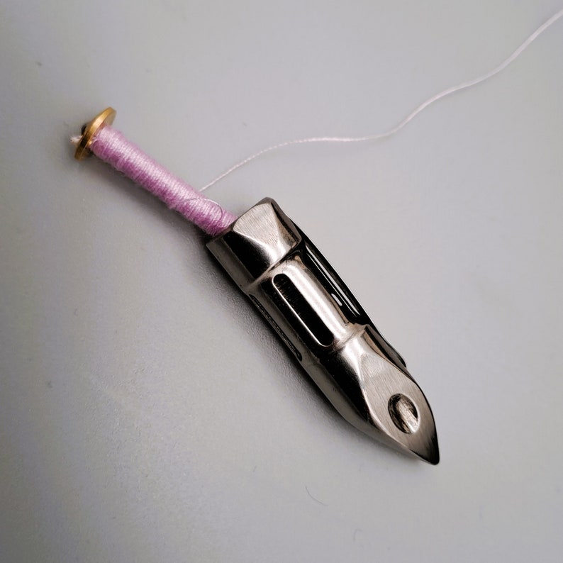 May include: A silver sewing machine bobbin winder with a pink thread wound around it. The thread is attached to a white thread.