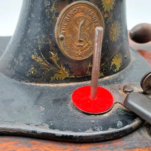 May include: Close-up of a vintage Singer sewing machine. The machine is black with gold floral accents and a brass emblem that reads "The Singer Mfg. Co." A red felt circle is visible.