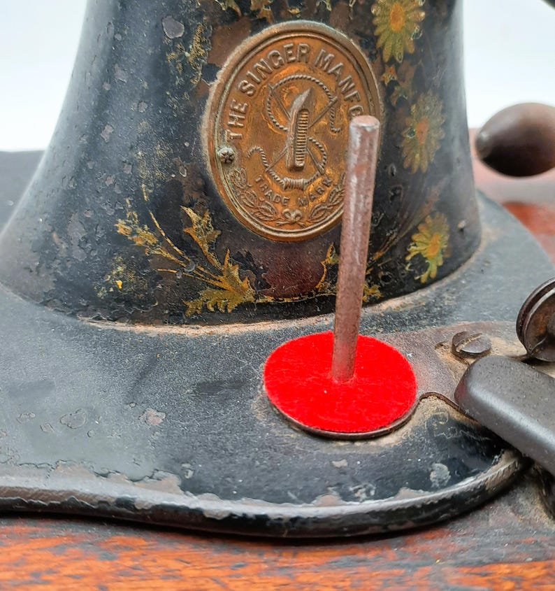 May include: Close-up of a vintage Singer sewing machine. The machine is black with gold floral accents and a brass emblem that reads "The Singer Mfg. Co." A red felt circle is visible.