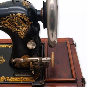 May include: Antique black sewing machine with gold floral detailing. The machine features a large silver flywheel and is set on a wooden base. A vintage sewing machine, perfect for collectors or sewing enthusiasts.