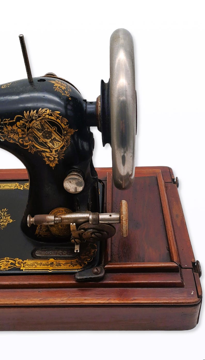 May include: Antique black sewing machine with gold floral detailing. The machine features a large silver flywheel and is set on a wooden base. A vintage sewing machine, perfect for collectors or sewing enthusiasts.