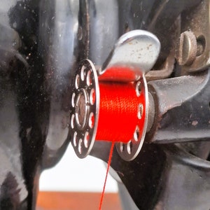 May include: Close-up of a sewing machine with a red thread spool and a metal thread guide. The thread is threaded through the guide and is ready to be used.