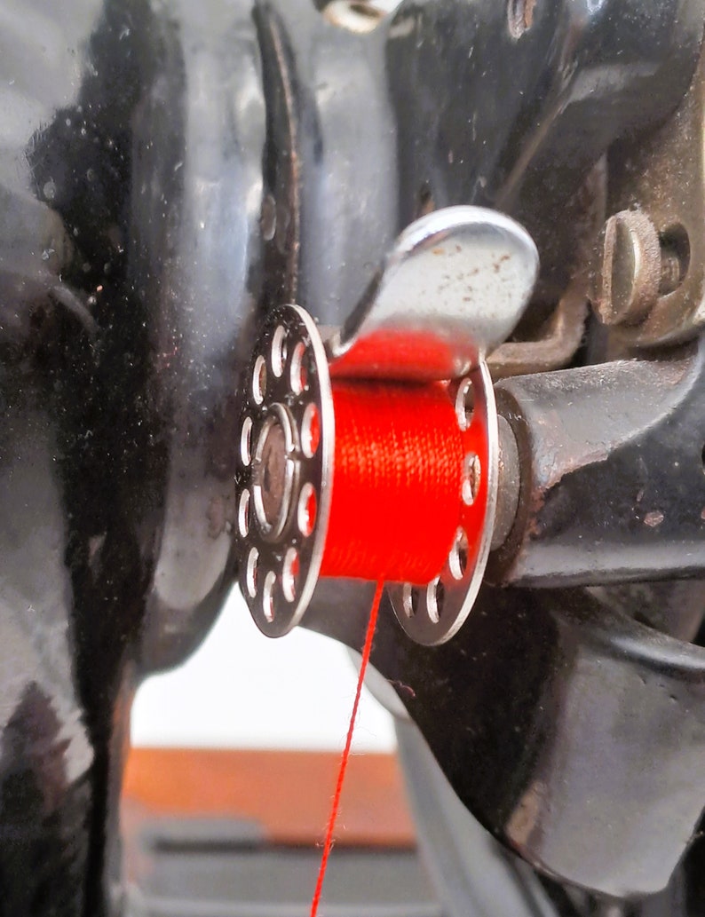 May include: Close-up of a sewing machine with a red thread spool and a metal thread guide. The thread is threaded through the guide and is ready to be used.