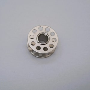 May include: A silver metal bobbin with a circular opening in the center and a series of small holes around the edge.