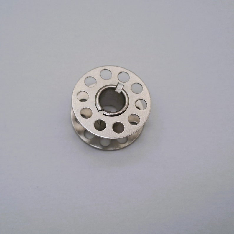 May include: A silver metal bobbin with a circular opening in the center and a series of small holes around the edge.