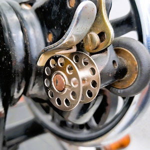 May include: Close-up of a vintage sewing machine with a silver metal spool holder and a black metal base.