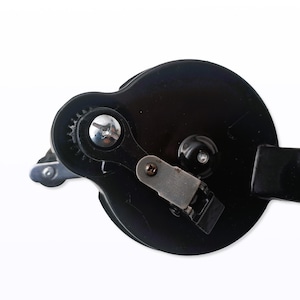 May include: Close-up of a black can opener with a silver gear and lever. The can opener has a circular design with a handle on the right side. The image is set against a white background.