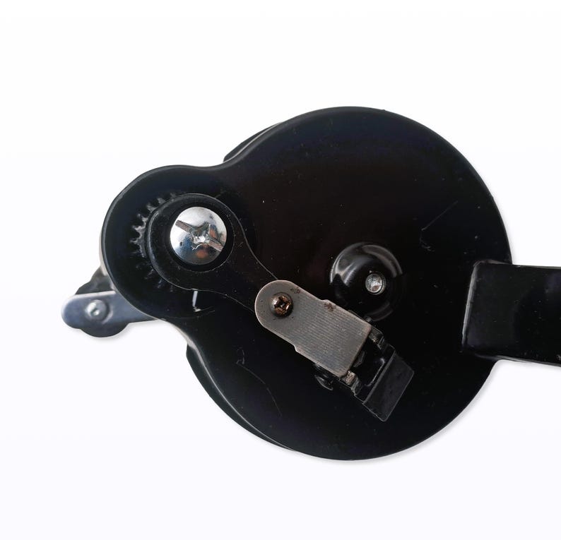 May include: Close-up of a black can opener with a silver gear and lever. The can opener has a circular design with a handle on the right side. The image is set against a white background.