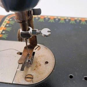 May include: Close-up of an antique sewing machine. The image highlights the needle, presser foot, and thread guide. The machine has a black body with decorative green and orange accents. The metal components show signs of age and use.