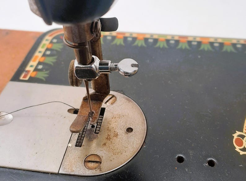 May include: Close-up of an antique sewing machine. The image highlights the needle, presser foot, and thread guide. The machine has a black body with decorative green and orange accents. The metal components show signs of age and use.