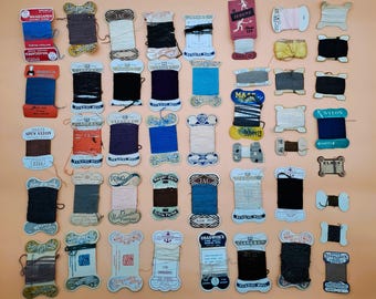 Könnte beinhalten: Eine Sammlung von Vintage-Garnrollen in verschiedenen Farben, darunter Blau, Schwarz, Braun und Weiß. Die Rollen sind auf einer pfirsichfarbenen Oberfläche angeordnet, wobei einige Etiketten sichtbar sind.