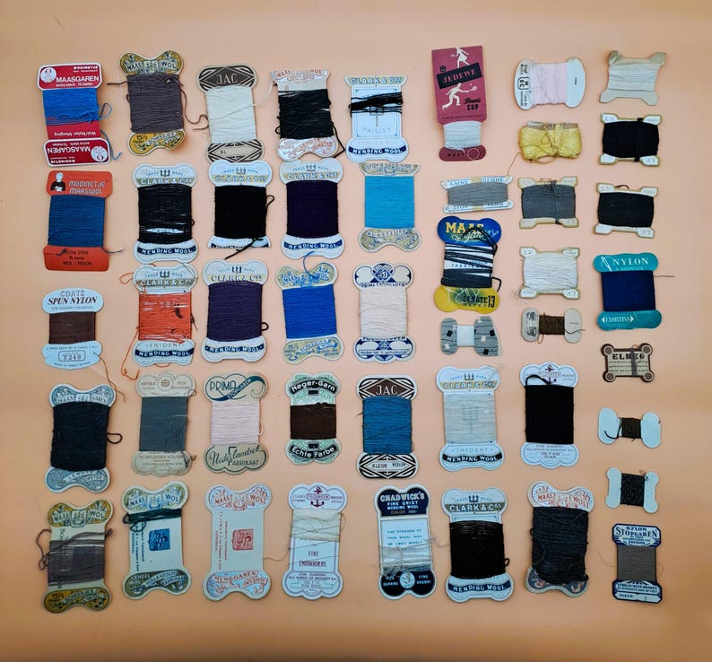 May include: A collection of vintage spools of thread in various colors, including blue, black, brown, and white. The spools are arranged on a peach-colored surface, with some labels visible.