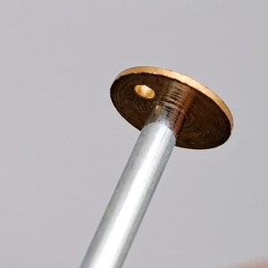 May include: A close-up of a brass-colored metal disc with a small hole in the center. The disc is attached to a silver metal rod.