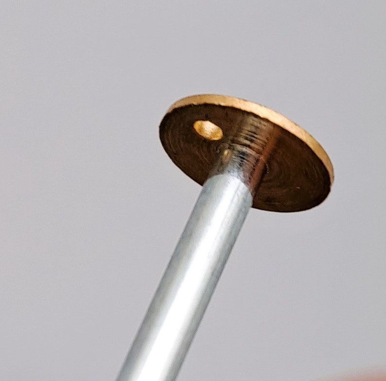 May include: A close-up of a brass-colored metal disc with a small hole in the center. The disc is attached to a silver metal rod.
