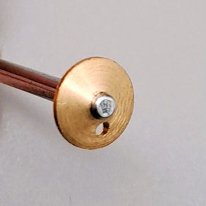 May include: A gold-colored metal watch part with a small hole and a crown logo on the top.
