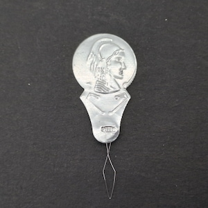 May include: Silver metal needle threader with a round head featuring a raised image of a woman's profile wearing a helmet. The threader has a small, pointed needle attached to the bottom.