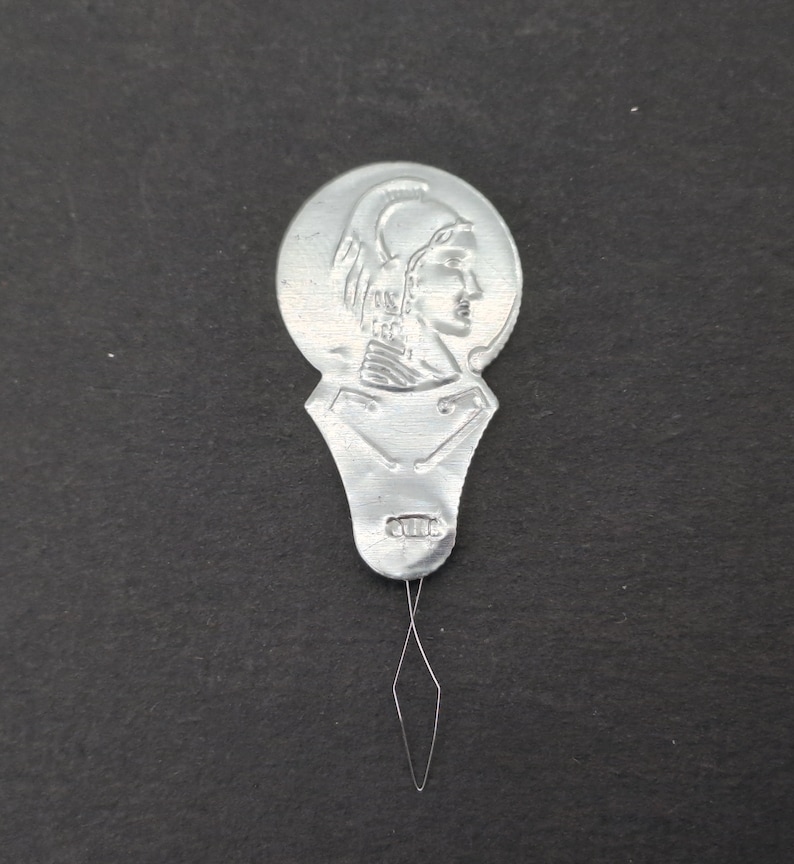 May include: Silver metal needle threader with a round head featuring a raised image of a woman's profile wearing a helmet. The threader has a small, pointed needle attached to the bottom.