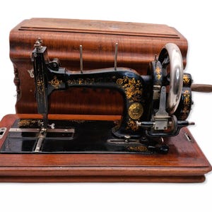 Könnte beinhalten: Eine antike schwarze Nähmaschine mit goldenen floralen Details, auf einem Holzsockel. Die Maschine hat ein silbernes Handrad und eine Holzabdeckung. Die Nähmaschine ist ein Vintage-Artikel.